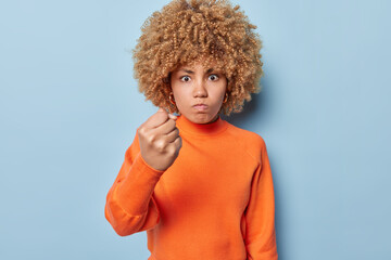 Outraged curly haired woman shaking clenched fist at camera showing her protest threatening someone expressing anger wears casual orange jumper isolated over blue background. Negative emotions