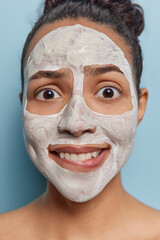 Close up image of worried Latin woman with dark hair looks nervously at camera bites lips applies white beauty mask undergoes skin care procedures at home poses against blue background in bathroom