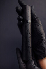 A mockup of a black tube for cream with drops of water. Close-up of the beautician hands in black gloves holding a moisturizer on a dark isolated background.