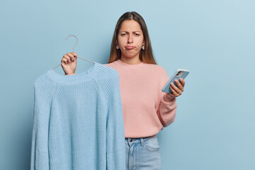 Photo of discontent European woman holds jumper on hanger uses mobile phone for surfing internet purses lips being dissatisfied with her purchase isolated over blue background. Shopping concept - obrazy, fototapety, plakaty