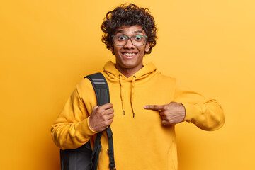 Positive curly haired Hindu man points at himself and being surprised being chosen asks who me carries rucksack on shoulder dressed in casual hoodie isolated over yellow background. Do you mean me