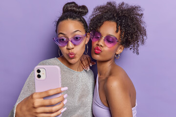 Two best female friends focused at camera of smartphone pouts lips and pose for selfie wear trendy sunglasses stylish clothing isolated over purple background. Friendship technology concept. - obrazy, fototapety, plakaty