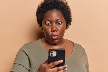 Photo of stupefied dark skinned woman checkes mailbox reads disturbing message received fascinating offer holds smartphone feels impressed wears casual jumper isolated over brown background. - obrazy, fototapety, plakaty