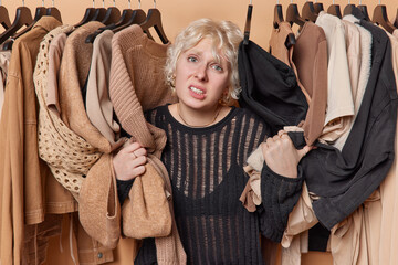 Confused hesitant young European woman clenches teth feels unaware poses among clothes on hangers...