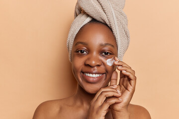 Natural beauty care. Cheerful dark skinned woman puts on facial cream wears bath towel on head...