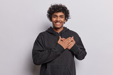 Waist up shot of happy Hindu man with curly hair keeps hands on heart expresses gratitude feels flattered and thankful smiles gladfully wears black sweatshirt isolated over white background.