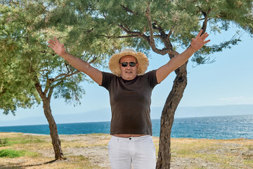 Overweight middle-aged smiling man wearing straw hat at seaside. Happy mature man with arms in up enjoying freedom at sea and looking at camera.