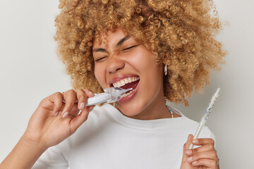 Photo of curly haired emotional woman holds plastic mouth expander and gel in syringe keeps mouth widely opened dressed in t shirt undergoes oral procedures isolated over white studio background