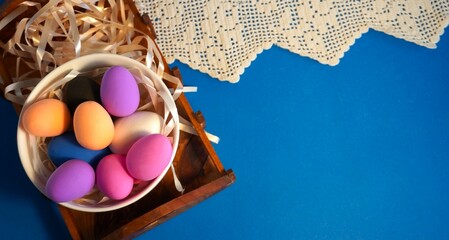 Easter composition of multicolored chicken eggs in a white porcelain plate on a wooden tray and a blue background with a vintage cotton lace napkin