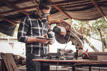 Male carpenter wearing protective mask using electric circular saw cutting wood board at workshop studio