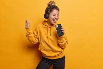 People emotions concept. Studio shot of young happy smiling excited European woman using headphones listening to music dancing holding smartphone as microphone singing wearing casual clothes