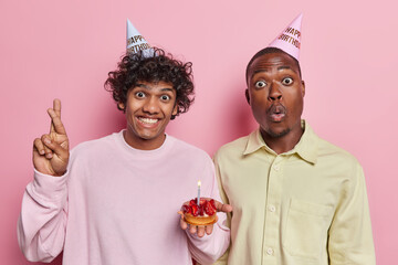 Studio photo of young happy smiling Hindu and surprised African american men celebrating birthday having party holding cake with candle standing close to each other on pink background wearing hats - Powered by Adobe