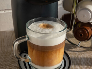Hot cappuccino coffee in a glass mug with top layer of frothy steamed milk foam and espresso on a coffee machine tray made from coffee capsules