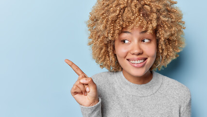 Studio shot of curly haired woman dressed in casual grey jumper points index finger on blank space for your advertisement isolated over blue background. People advertisement and promotion concept