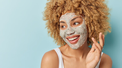 Beautiful cheerful curly haired woman applies facial clay mask for skin treatment focused aside...
