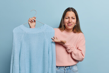 Displeased young woman with long hair holds knitted jumper on hanger being dissatisfied with...