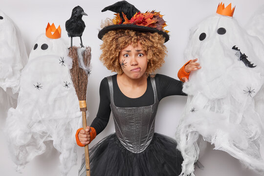 Organising halloween party. Indoor shot of young beautiful aggressive African american woman in which costume posing on white background surrounded by scary white ghosts with broom and raven on top