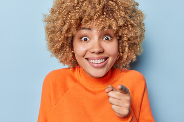 Positive curly haired woman with toothy smile points index finger at camera dressed in casual orange jumper isolated over blue background. Optimistic female model chooses someone. You should follow me