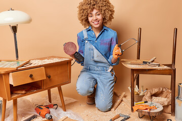 Professional curly haired female carpenter or labor worker holds electric using wood polishing machine grinding wears blue shirt and denim overalls produces furniture isolated over brown background