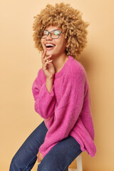 Vertical shot of overjoyed curly haired woman laughs joyfully as hears something funny poses on chair being in good mood wears spectacles pink jumper and jeans isolated over brown background