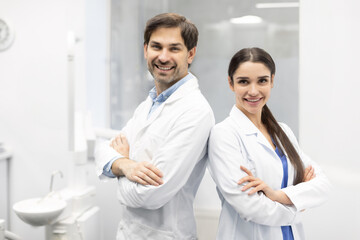 Smiling male doctor and female assistant with crossed hands looking at camera, posing in dental clinic interior