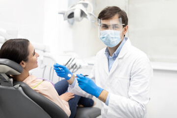 Male dentist smiling and working in dentist office, repairing teeth of young woman, dentist looking at camera wearing white uniform, mask and dentistry glasses