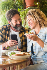 Adult couple smile and enjoy lunch and phone video call sitting at the bar and laughing together with happiness - people man and woman use cellular and have brunch in restaurant