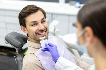 Female dentist applying tooth scale samples set to smiling male patient of dental clinic, closeup...