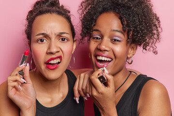 Horizontal shot of displeased Latin woman holds red lipstick poses with her best friend undergo...