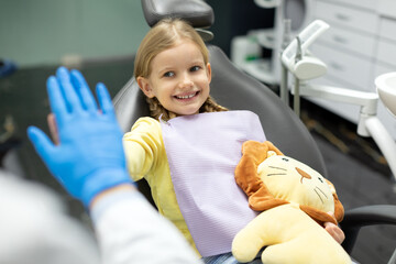 Children's dentistry. Smiling child girl sitting in the dental chair and cheerfully gives high five to male doctor