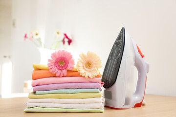 Stack of clean clothes, modern iron and flowers on wooden table