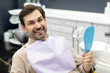 Portrait of happy male patient holding mirror after treatment and smiling, sitting in chair in stomatological cabinet