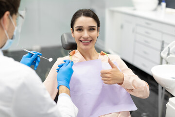 Happy female patient showing thumb up while having check up with male dentist in modern stomatologic clinic, enjoying quality dental treatment and medical service