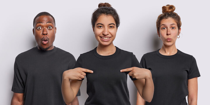Smiling Iranian woman points at mock up space of casual t shirt being in good mood stands in centre shocked African man and amazed female model stand against white background. Collage image.