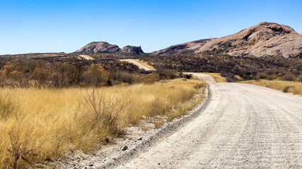 Erongo mountains, Namibia - August 16, 2022: A winding gravel road stretches into the distance, flanked by golden grasses and leading towards rugged mountains under a clear blue sky.