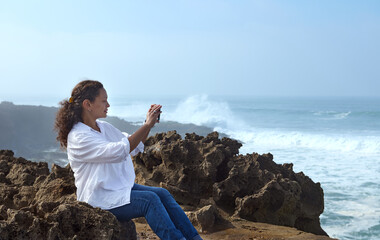 Naklejka na ściany i meble Side view of a happy relaxed woman sitting on the cliff and photographing the ocean on her smartphone