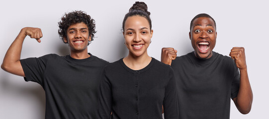 Horizontal shot of pleased Latin woman with hair bun poses among two men who clench fists from triumph and show biceps dressed in black t shirts isolated over white background. Indoor studio image
