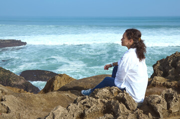 Beautiful woman dreamily looking into the distance, sitting on the cliff by ocean shore, feeling connection with nature