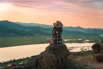 hatted woman in checkered shirt admiring landscape of mountains and river during the sunset. the...