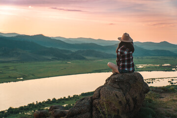hatted woman in checkered shirt on the top of the mountain in summer against the sunset over the...
