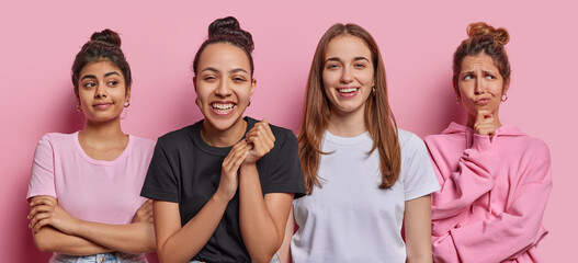 Four diverse women smile gladfully stand next to each other think about something deeply isolated over pink background express different emotions. Two happy female models between thoughtful.