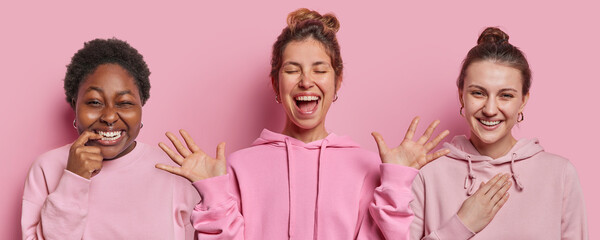 Photo of cheerful mixed race women smile happily exclaim from joy keep palms raised up react to something funny dressed in casual sweatshirt isolated over pink background. Multiracial female models