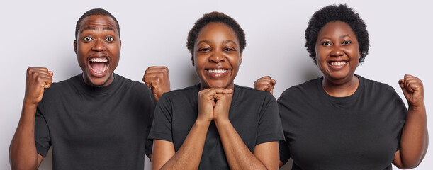 Image of positive African woman keeps hands under chin smiles broadly poses between triumphing female and male dressed in casual black t shirts isolated over white background. Three emotional people