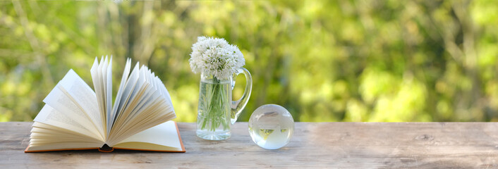 paper book, glass ball globe, bouquet of wild garlic flowers on old wooden table in garden, blurred...