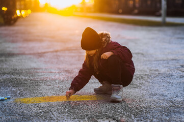Little diligent lonely girl, child draws a drawing with colored chalk on the asphalt outdoors on the street in the park at sunset. Photography, close-up portrait, design, childhood concept.
