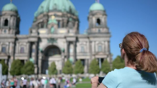 Berlin,Germany,August 10,2023. A middle-aged woman is standing  in front of the cathedral,taking photos with her cell phone,in the blurred background other people and the facade. Beautiful summer day.