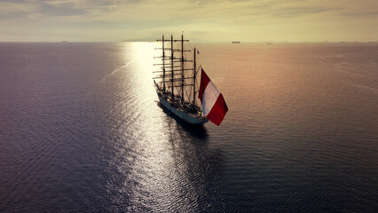 Aerial drone photo of beautiful three mast barque or barc type classic sailing wooden boat with huge waving flag anchored in Mediterranean deep blue Aegean sea at sunset with golden colours