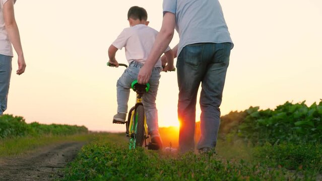 Father holds bicycle seat supporting child pedaling near walking mother. Happy mother looks at father teaching child riding bicycle. Father controls child riding bike with mother nearby at sunset