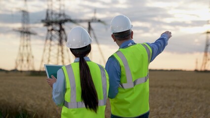 Electricians with tablet computer walk past power generation substation at countryside. Engineers...