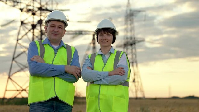 Cheerful electricians colleagues stand crossing arms against electric power transmission lines in sunset field. Engineers team with crossed arms rest after work at power distribution substation